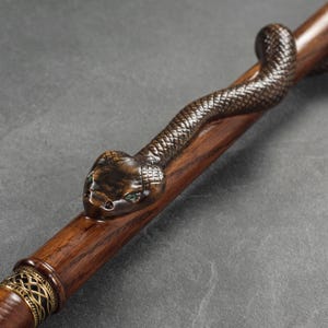 May include: A wooden walking stick with a carved snake head at the top. The snake has green eyes and a brown and black body.