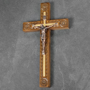 May include: A wooden cross with a carved floral design and a bronze-colored figure of Jesus Christ. The cross has a gold-colored inlay and is mounted on a gray wall. The cross is a religious item.
