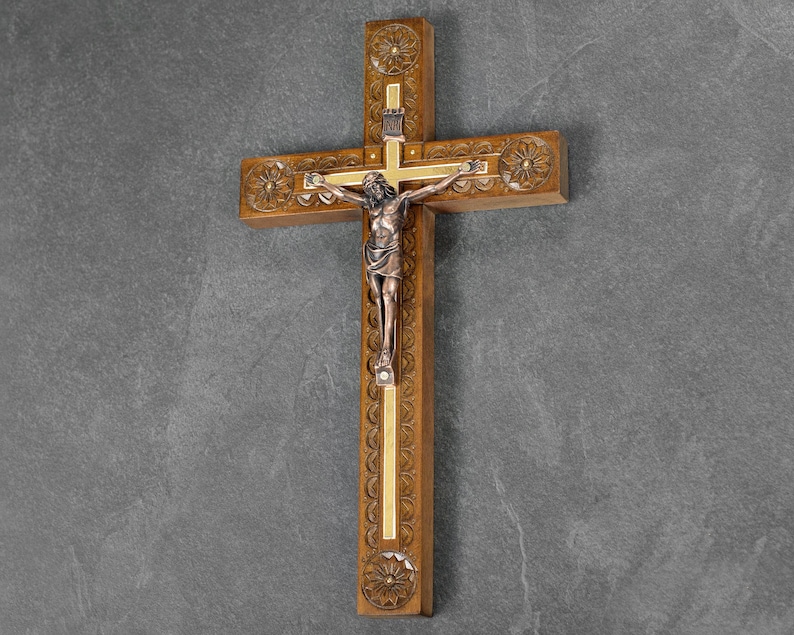 May include: A wooden cross with a carved floral design and a bronze-colored figure of Jesus Christ. The cross has a gold-colored inlay and is mounted on a gray wall. The cross is a religious item.