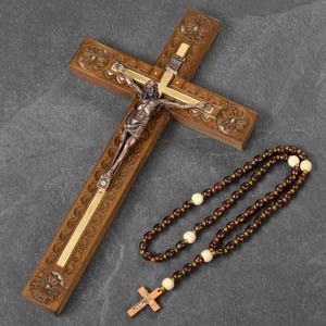 May include: A wooden crucifix with a bronze figure of Jesus Christ and a rosary. The cross is carved with floral designs and has a gold-colored inlay. The rosary features dark brown beads and cream-colored accent beads.