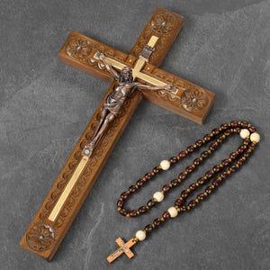 May include: A wooden crucifix with a bronze Jesus figure and a rosary. The cross is brown with carved floral designs and a gold-colored inlay. The rosary has brown beads with white accents and a small wooden cross.