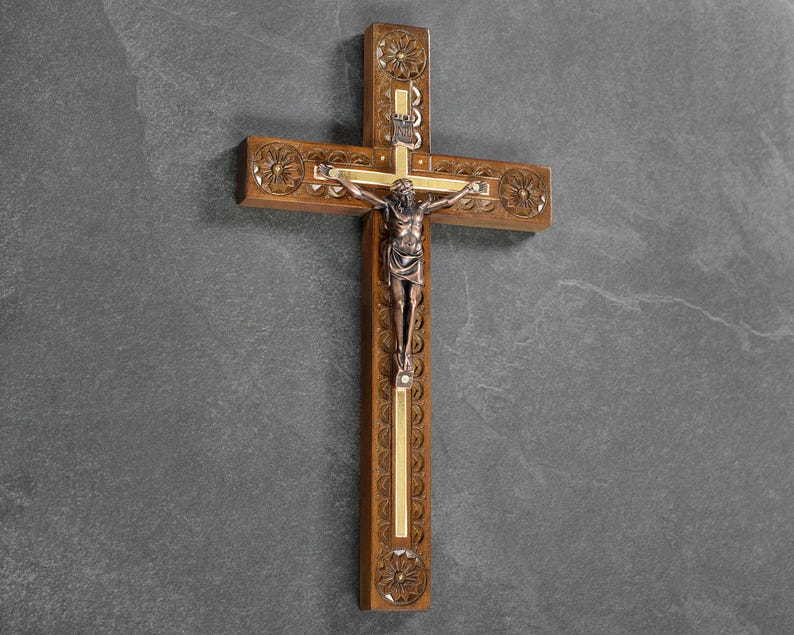 May include: A wooden crucifix with a detailed carving and a bronze figure of Jesus. The cross features decorative floral patterns and gold accents. The figure is centrally positioned on the cross.