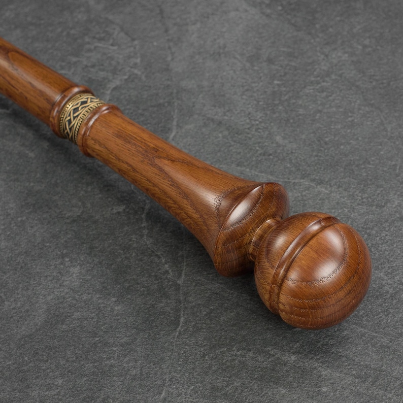 May include: A wooden walking stick with a rounded knob handle. The stick is brown and has a gold band with a decorative pattern.
