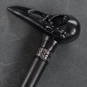 May include: A black walking stick with a detailed crow skull handle and a silver band with the words "Handmade" and "Este.com" engraved on it.