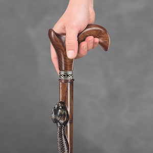 May include: A wooden walking stick with a snake head carved into the bottom of the shaft. The handle is a dark brown colour and has a decorative silver band.