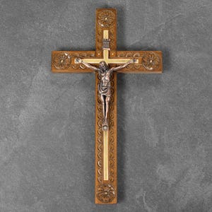 May include: Wooden crucifix with a detailed carved design. The cross features a silver-toned figure of Jesus Christ. The cross is made of brown wood with gold accents and decorative floral carvings.