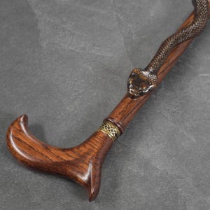 May include: A wooden walking stick with a carved snake head at the top. The stick is brown and has a gold band around it.