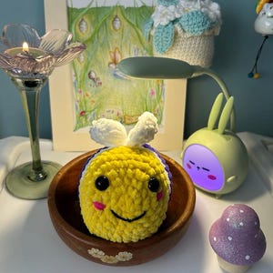 May include: A crocheted bee toy in a wooden bowl. The bee is yellow with purple stripes and white antennae. A small purple mushroom and a green lamp are also visible. The scene is set indoors.