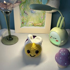 May include: A crocheted bee toy with a yellow body, white and purple details, and a smiling face. A bunny-shaped night light and a purple glitter mushroom are also in the image. A framed picture and a candle holder are also visible.