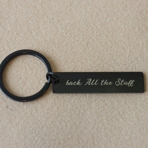 以下が含まれることがあります： 「back all the stuff」と書かれた長方形のタグが付いた黒い金属製のキーホルダーです。