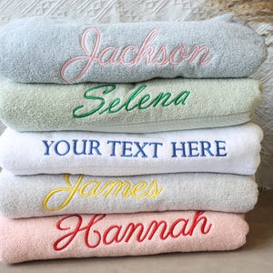 May include: A stack of five towels with different colored embroidery. The top towel is light blue with pink embroidery that says "Jackson". The second towel is light green with green embroidery that says "Selena". The third towel is white with blue embroidery that says "YOUR TEXT HERE". The fourth towel is light gray with yellow embroidery that says "James". The bottom towel is pink with red embroidery that says "Hannah".