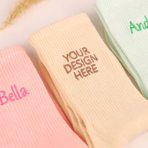 Könnte beinhalten: Nahaufnahme von mehreren Paar pastellfarbenen Socken. Ein Paar ist rosa mit "Bella" in Rosa gestickt, ein anderes ist cremefarben mit "YOUR DESIGN HERE" in Braun gestickt, und ein drittes ist mintgrün mit "Andy" in Grün gestickt.