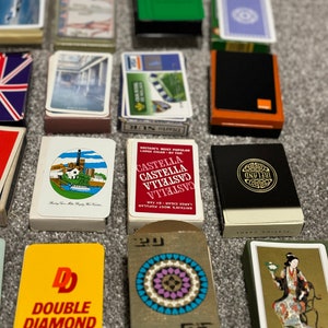 May include: A collection of various playing card decks, including a deck with a double diamond logo, a deck with a colorful circular design, and a deck with a black and gold design featuring the word "Ireland".