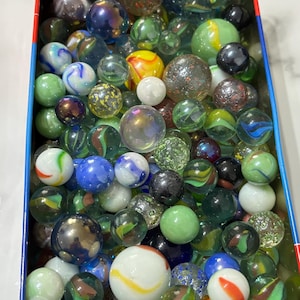 Peut inclure: Une boîte en étain remplie de billes colorées. Les billes sont en verre et présentent différents motifs et couleurs, notamment transparent, vert, bleu, rouge et blanc.