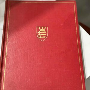 May include: A red book with a gold border and a gold emblem. The book has a textured cover and is likely a hardcover edition. The emblem is a shield shape with a stylized design.