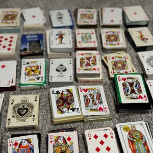 May include: A collection of playing cards, including standard decks and single cards, with various designs and colors. Some cards feature traditional designs, while others have unique illustrations. The cards are arranged on a gray surface.