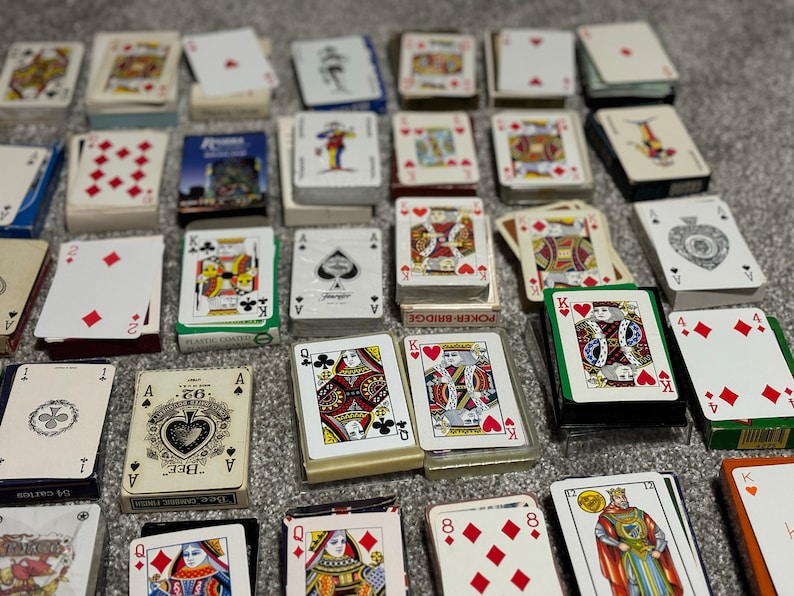 May include: A collection of playing cards, including standard decks and single cards, with various designs and colors. Some cards feature traditional designs, while others have unique illustrations. The cards are arranged on a gray surface.