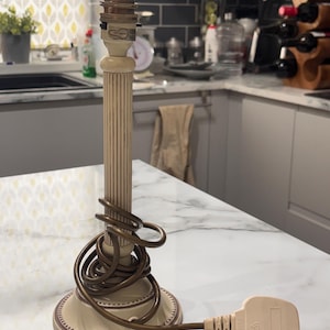 May include: A cream-coloured lamp base with a fluted column design and a round, decorative base. A brown electrical cord is wrapped around the column, ending in a standard three-pin plug. The top features a brass-coloured metal light socket.