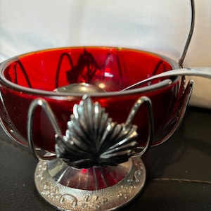 May include: A red glass sugar bowl with a silver leaf design and a silver wire handle. The bowl is sitting on a silver base with a floral design.