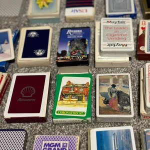 May include: A collection of vintage playing card decks featuring various designs, including a red deck with a Shell logo, a green deck with a pub scene, a blue deck with a winter scene, a purple deck with the MGM Grand logo, and a blue deck with an airplane. The decks are arranged on a brown surface.