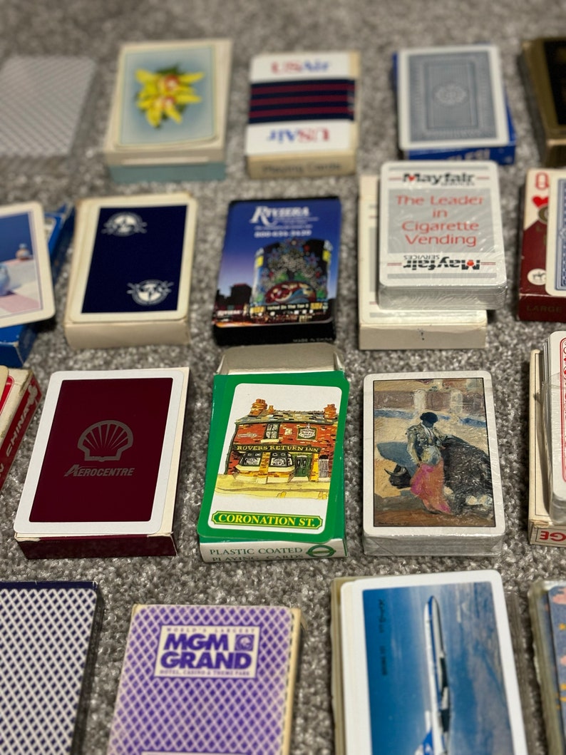 May include: A collection of vintage playing card decks featuring various designs, including a red deck with a Shell logo, a green deck with a pub scene, a blue deck with a winter scene, a purple deck with the MGM Grand logo, and a blue deck with an airplane. The decks are arranged on a brown surface.