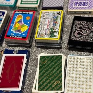 May include: A collection of playing card decks with various designs. Some decks feature a map of Barbados, a map of Vancouver, and a green deck with a Carlsberg logo. Other decks have a white background with a repeating pattern of circles or triangles.
