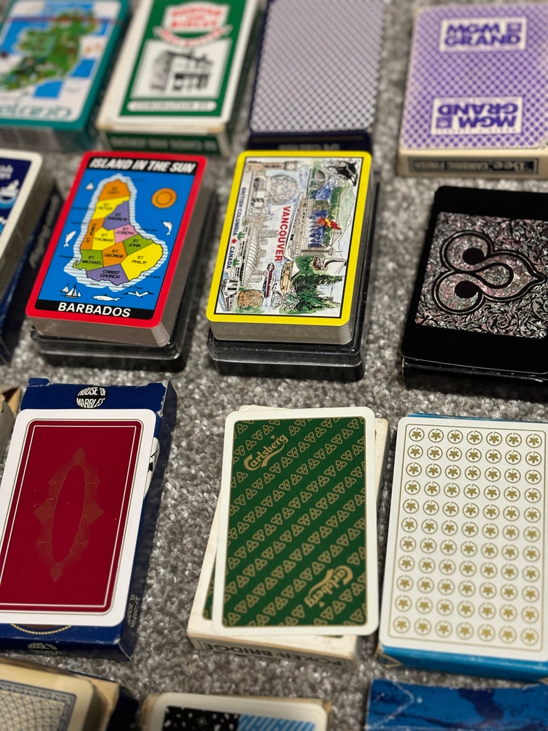 May include: A collection of playing card decks with various designs. Some decks feature a map of Barbados, a map of Vancouver, and a green deck with a Carlsberg logo. Other decks have a white background with a repeating pattern of circles or triangles.
