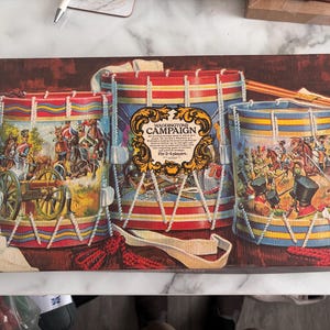 May include: A rectangular board game box titled Waddingtons Campaign for 2-4 players. The box features an illustration of three drums decorated with historical military scenes, including soldiers on horseback and cannons, with white rope lacing.