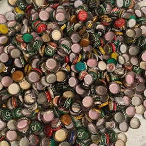 May include: A collection of mixed metal bottle caps, displaying a range of colours and designs. The caps are scattered, with visible text and logos. The assortment seems to be from different brands, creating a diverse mix.