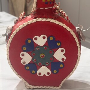 May include: A round red canteen featuring a decorative pattern of white and blue hearts surrounding a central dark teal floral motif. The edges are trimmed with braided white leather cord, and the top includes a matching red leather stopper.