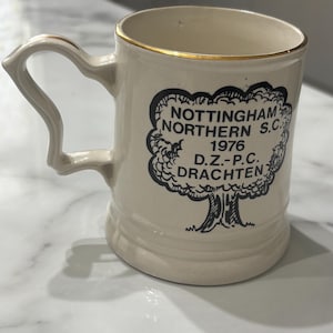 May include: A cream-coloured ceramic mug with a gold-trimmed rim and a decorative handle. The front features a black ink illustration of a tree with the text: NOTTINGHAM NORTHERN S.C. 1976 D.Z.-P.C. DRACHTEN printed in the centre of the tree canopy.