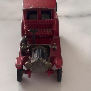 May include: A vintage red die-cast toy car featuring an open-top design, a black steering wheel, and a silver-toned radiator grille. The vehicle has small black wheels, a textured red bench seat, and visible paint wear along the edges of the roof.