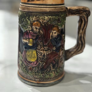 May include: A ceramic beer stein featuring a relief design of two figures in a forest setting. The mug has a textured handle resembling wood grain, a tan rim, and a dark base. The figures are painted in shades of red, yellow, and blue.