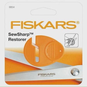 May include: An orange Fiskars SewSharp Restorer tool in its packaging. The package has the Fiskars logo and the text "SewSharp Restorer". The tool is designed to sharpen scissors. The packaging is white and orange.