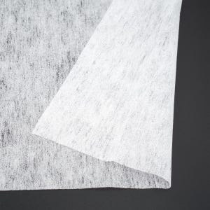 Pode incluir: Close-up de um tecido branco não tecido com uma superfície ligeiramente texturizada. O tecido está dobrado, revelando uma aparência em camadas. O fundo é preto sólido.