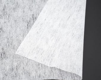Op de afbeelding: Close-up van een witte, non-woven stof met een licht getextureerd oppervlak. De stof is dubbelgevouwen, waardoor een gelaagde uitstraling ontstaat. De achtergrond is effen zwart.