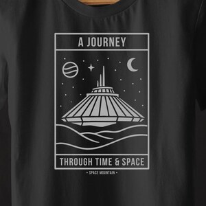 以下が含まれることがあります： スペースマウンテンのデザイン、惑星、三日月が描かれた白いグラフィックの黒いTシャツ。Tシャツのテキストは「A Journey Through Time & Space - Space Mountain」と書かれています。Tシャツは木製のハンガーにかかっています。
