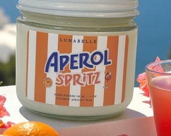 May include: A glass jar candle with a white lid and a label that reads "Aperol Spritz." The label has orange and white stripes and a graphic of an Aperol Spritz cocktail. The candle is surrounded by oranges and pink flowers.