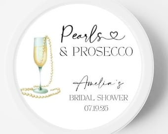 May include: A round white sticker featuring a watercolor illustration of a champagne flute draped with a string of pearls. The text reads Pearls & PROSECCO, Amelia's BRIDAL SHOWER, and the date 07.19.25 in black script and serif fonts.