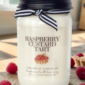 May include: A glass jar candle with a black lid and a black and white striped ribbon. The label reads "RASPBERRY CUSTARD TART" with a raspberry tart illustration. The candle is 16 oz.
