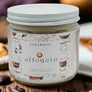 May include: A white candle jar with a white lid. The jar has a label that says "LUNABELLE affogato" and has a hand-drawn illustration of a coffee drink with whipped cream. The label also says "HAND-POURED IN NJ | 1.12 OZ COCONUT APRICOT WAX". The jar is sitting on a wooden surface with coffee beans and a pastry.