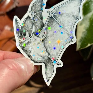 May include: A die-cut sticker featuring a detailed black and white ink illustration of a bat in flight. The bat has small red eyes and is overlaid with a holographic star pattern that reflects light in various colors across its wings and body.