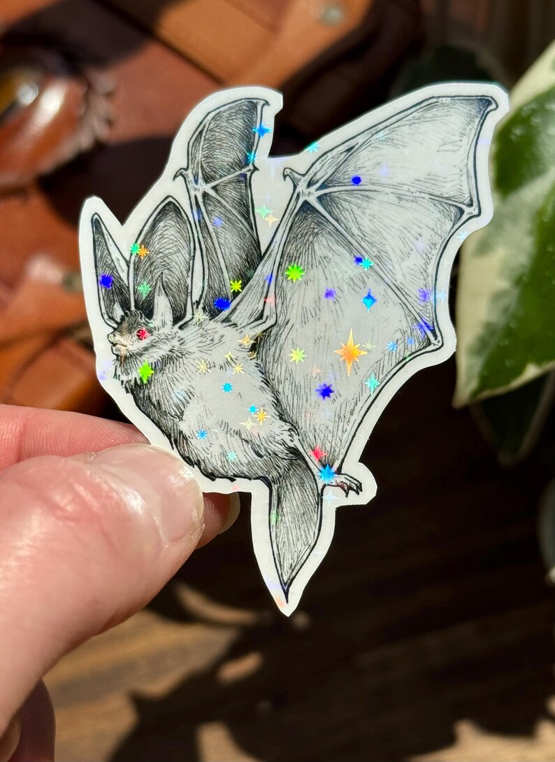 May include: A die-cut sticker featuring a detailed black and white ink illustration of a bat in flight. The bat has small red eyes and is overlaid with a holographic star pattern that reflects light in various colors across its wings and body.