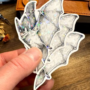 May include: A die-cut sticker featuring a detailed black and white ink illustration of a bat in flight. The sticker has a holographic finish that creates a shimmering star-like effect across the wings and body of the bat, highlighting the fine lines.