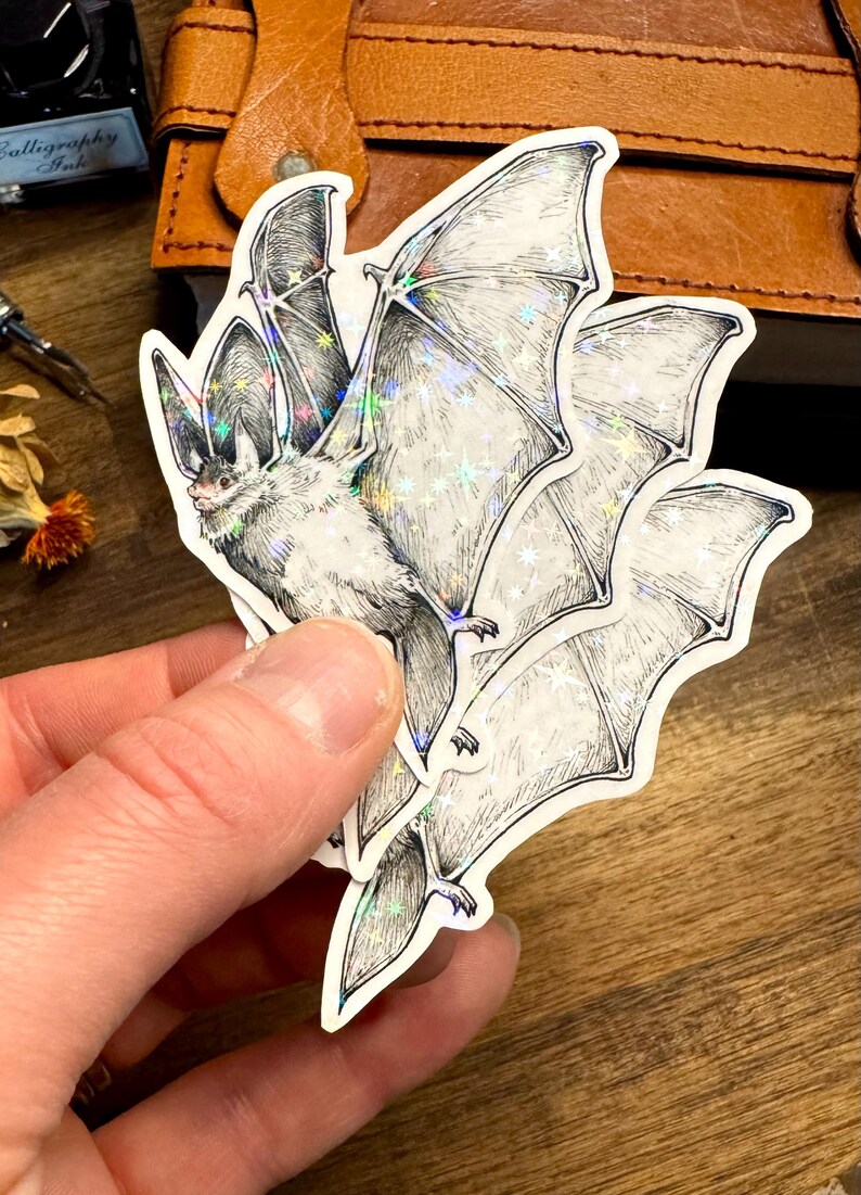 May include: A die-cut sticker featuring a detailed black and white ink illustration of a bat in flight. The sticker has a holographic finish that creates a shimmering star-like effect across the wings and body of the bat, highlighting the fine lines.