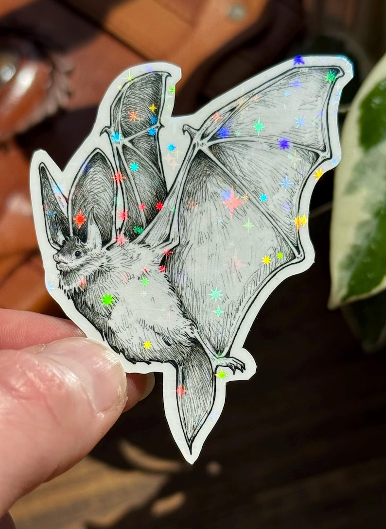 May include: A die-cut sticker featuring a black and white ink illustration of a bat in flight with its wings spread wide. The sticker surface has a holographic finish with small, colorful star-shaped accents scattered across the bat's body and wings.