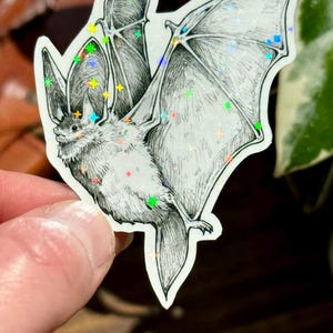 May include: A die-cut sticker featuring a detailed black and white ink illustration of a bat in flight. The bat is depicted with its wings spread wide, and the surface of the sticker is covered in small, holographic, multi-colored star accents.