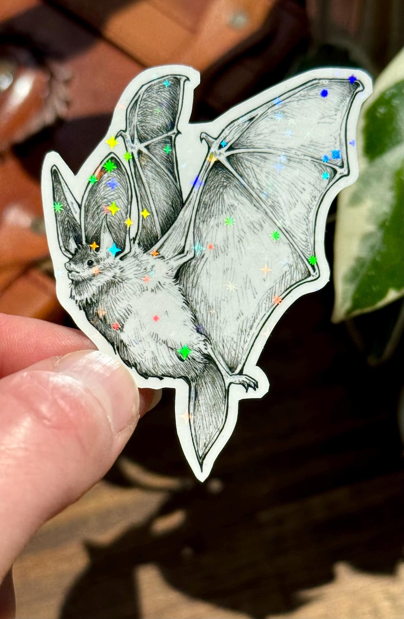 May include: A die-cut sticker featuring a detailed black and white ink illustration of a bat in flight. The bat is depicted with its wings spread wide, and the surface of the sticker is covered in small, holographic, multi-colored star accents.