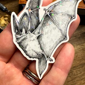 May include: A die-cut sticker featuring a detailed black and white ink illustration of a bat with large ears and outstretched wings. The sticker has a holographic finish that reflects light with small star-shaped sparkles across the wing surface.