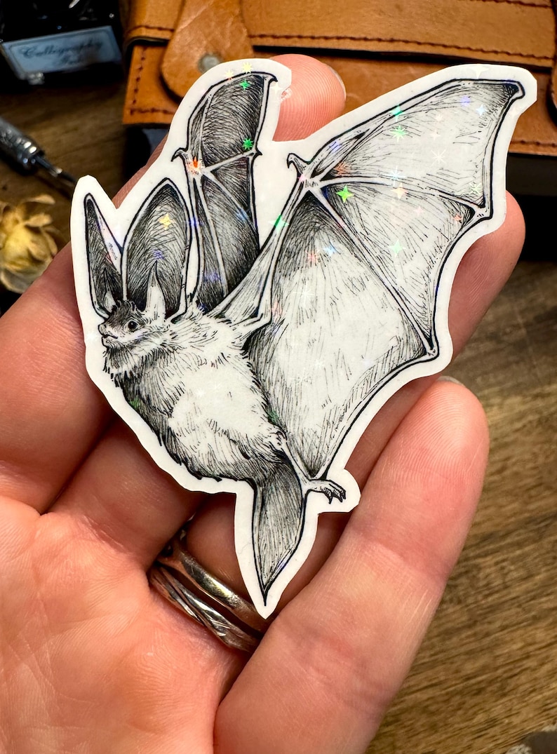 May include: A die-cut sticker featuring a detailed black and white ink illustration of a bat with large ears and outstretched wings. The sticker has a holographic finish that reflects light with small star-shaped sparkles across the wing surface.